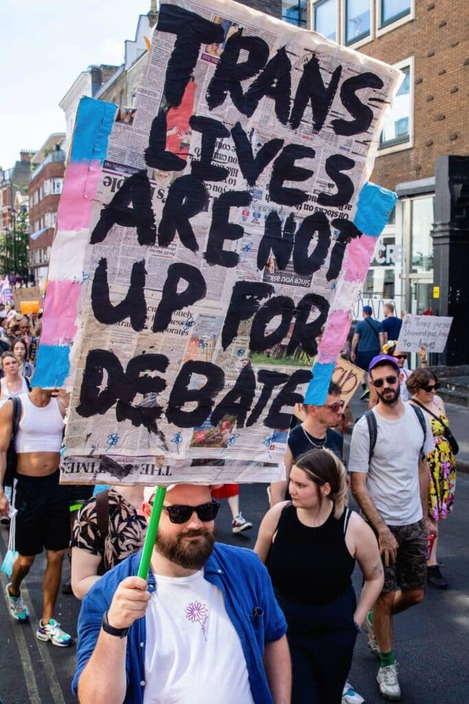 A person holds up a sign that reads 'Trans lives are not up for debate' with blue, pink and white stripes representing the trans pride flag on the side of the sign