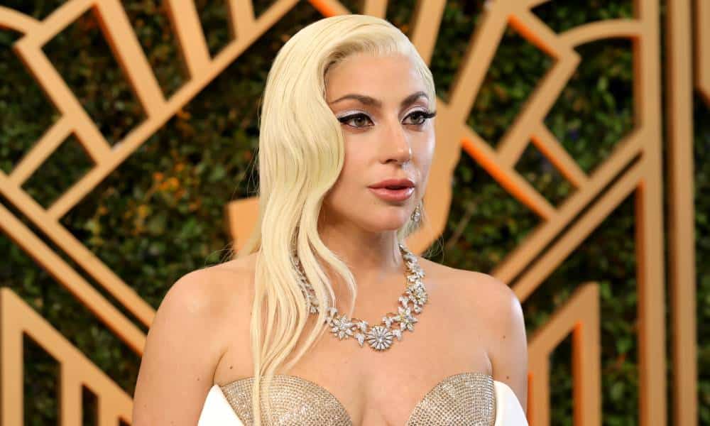 Lady Gaga wears a white dress with sparkly gold details as she stands in front of a gold and black background