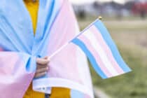 A trans person holding a flag with the trans flag draped over them.