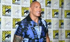 Dwayne 'The Rock' Johnson smiles for the camera as he wears a blue and black patterned top and stands in front of a patterned background with the SD Comic Con logo on it