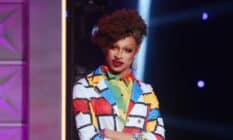 Drag Race star Yvie Oddly wears a white, blue, red and yellow geometric patterned outler layer with another colourful layer of clothing below. Yvie's hair is styled in an updo