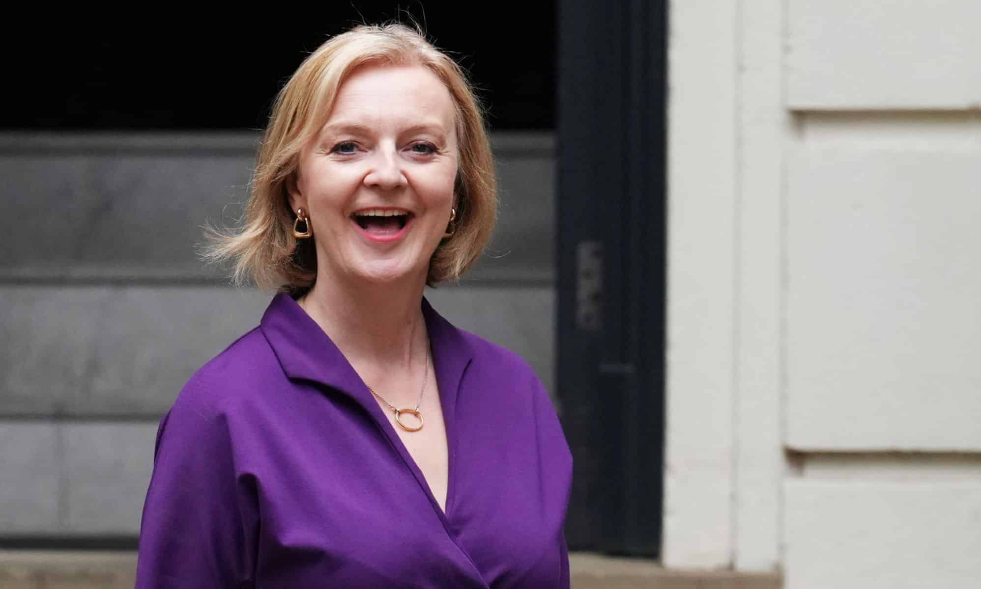Liz Truss sourit alors qu'elle se tient devant un immeuble portant une tenue violette