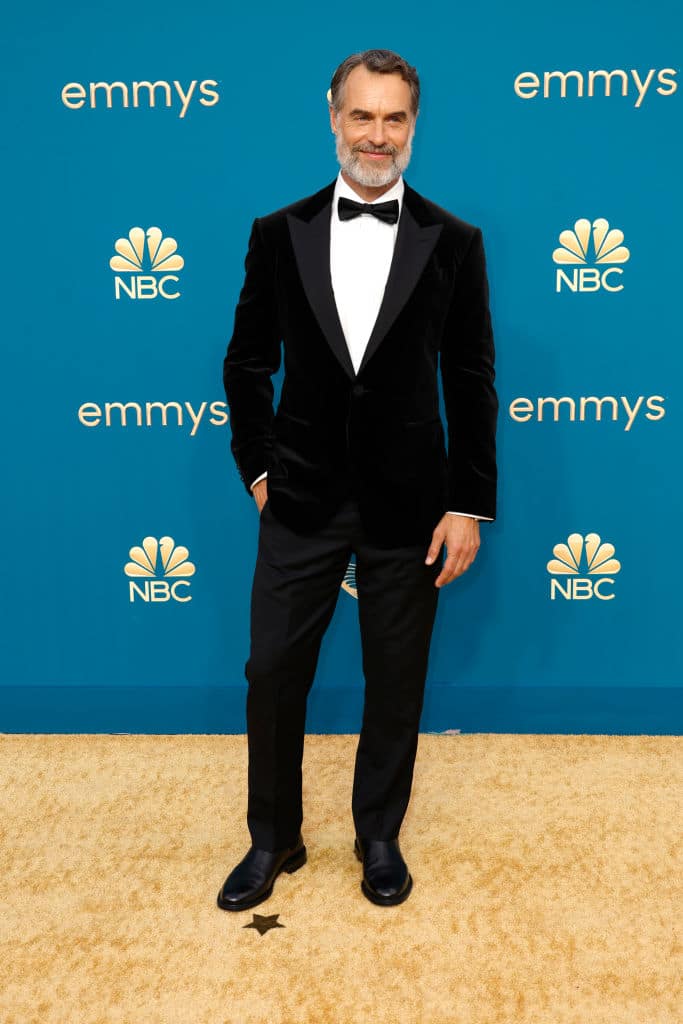 Murray, en costume noir et chemise blanche, assis contre un mur bleu sur un tapis doré