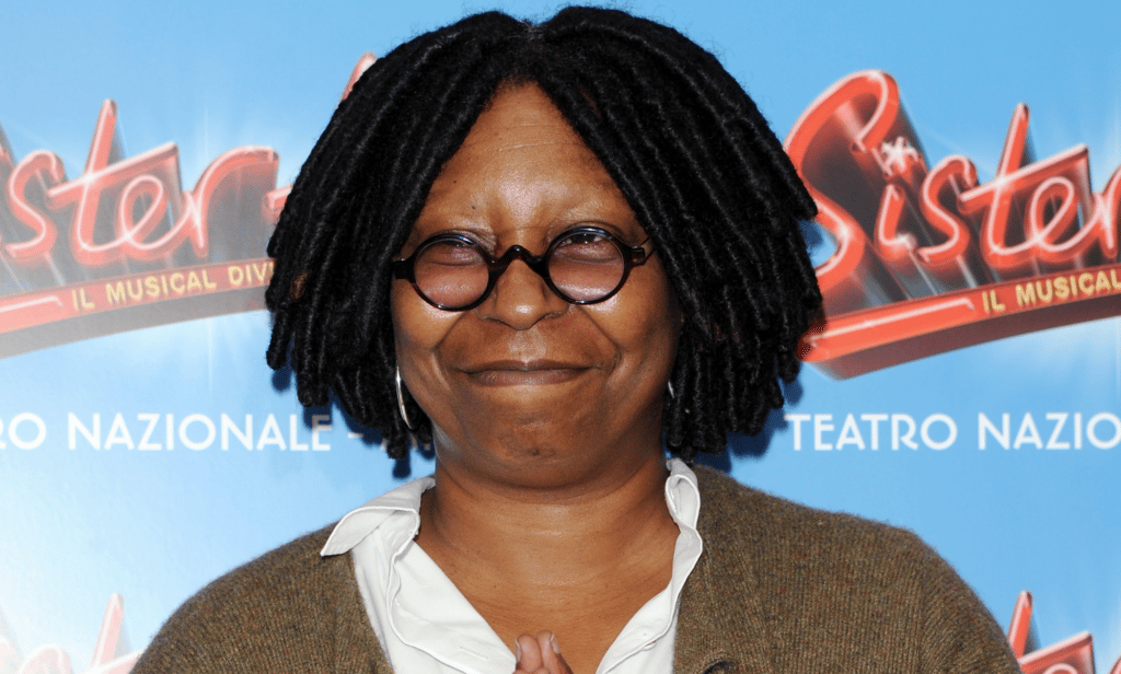 Whoopi Goldberg wears a white shirt and brown top over it as she smiles at the camera